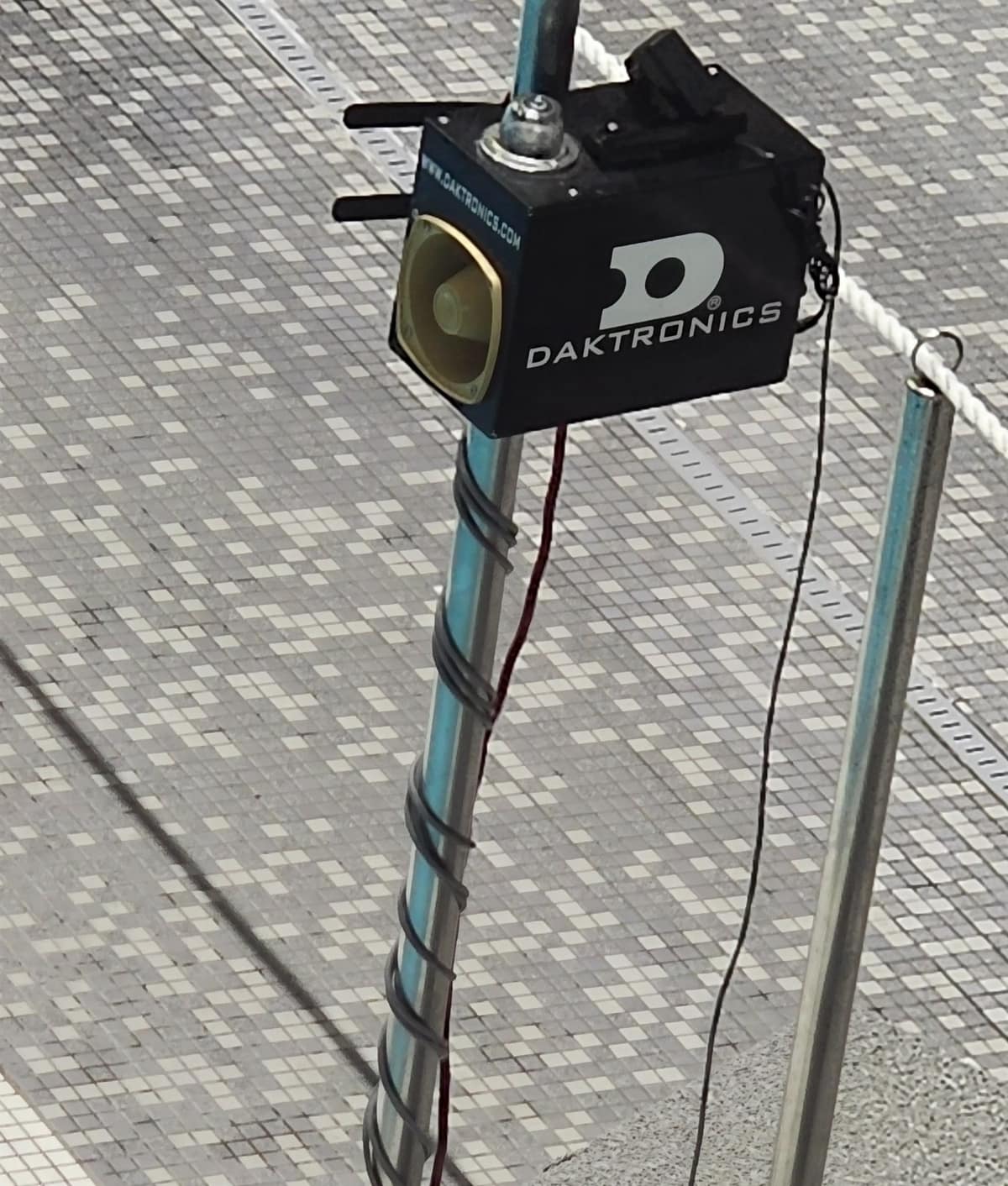 A black Daktronics unit clamped to the top of a backstroke flagpole on a tiled pool deck, with a round horn speaker in its front face and cables running down the pole.